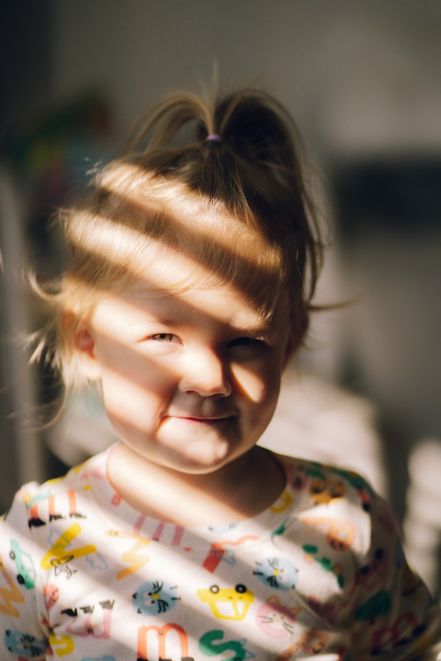 smiling toddler indoors with sunlight stripes across their face and colorful shirt