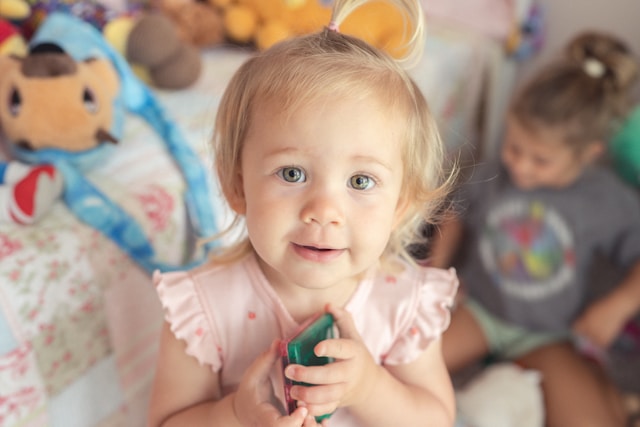 toddler looking up at the camera while holding a small toy, with another child and stuffed animals in the background
